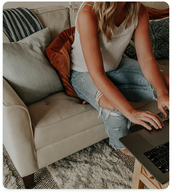 A woman typing on a desktop while sitting on a grey couch.